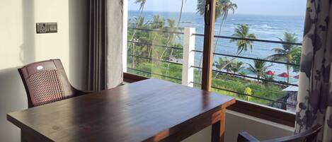 A balcony with a wooden table and chairs, overlooking a beach with palm trees and the ocean.