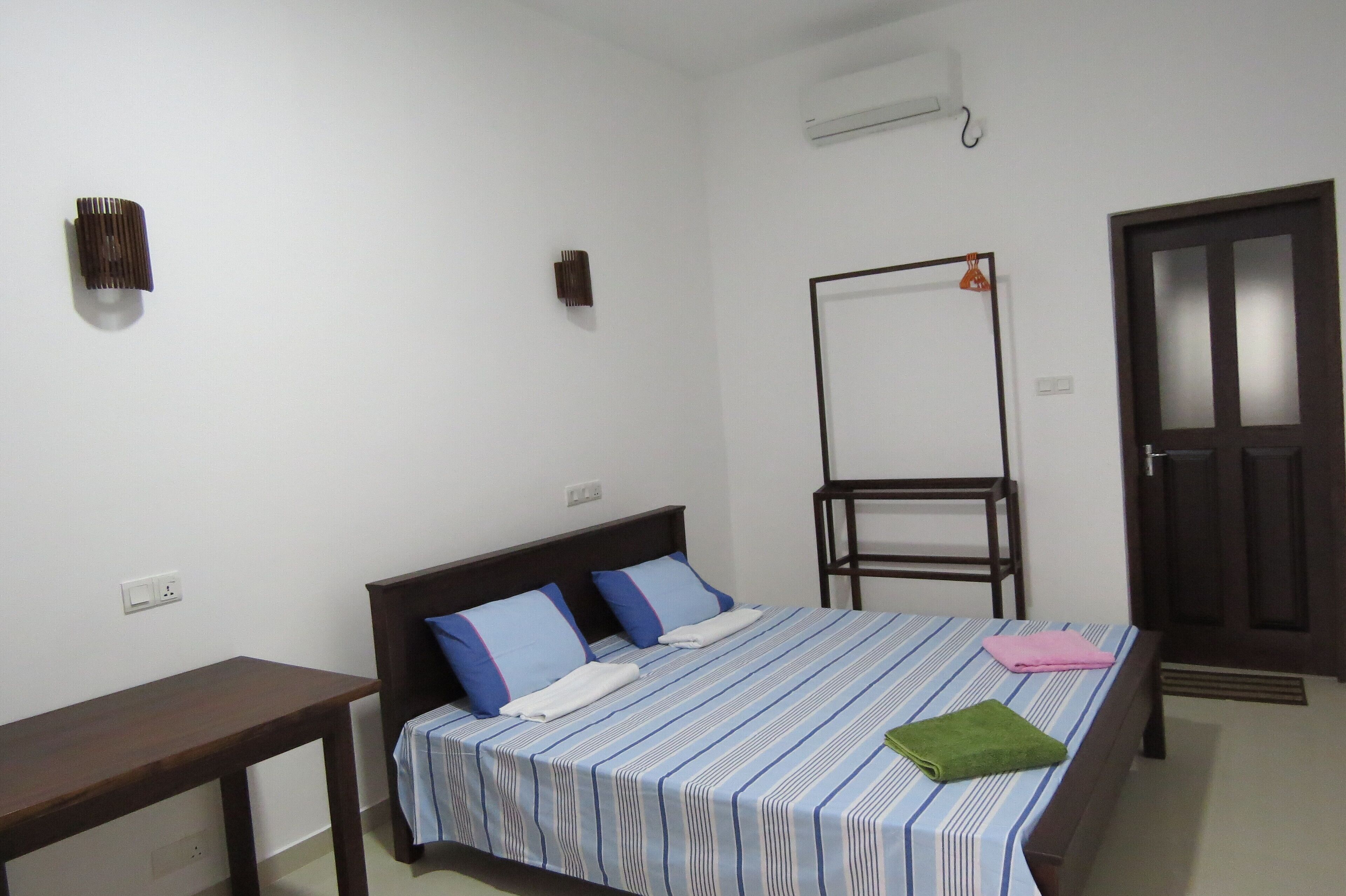 A single-bed room with a striped bedspread, two pillows, a bedside table, a standing fan, and a door.