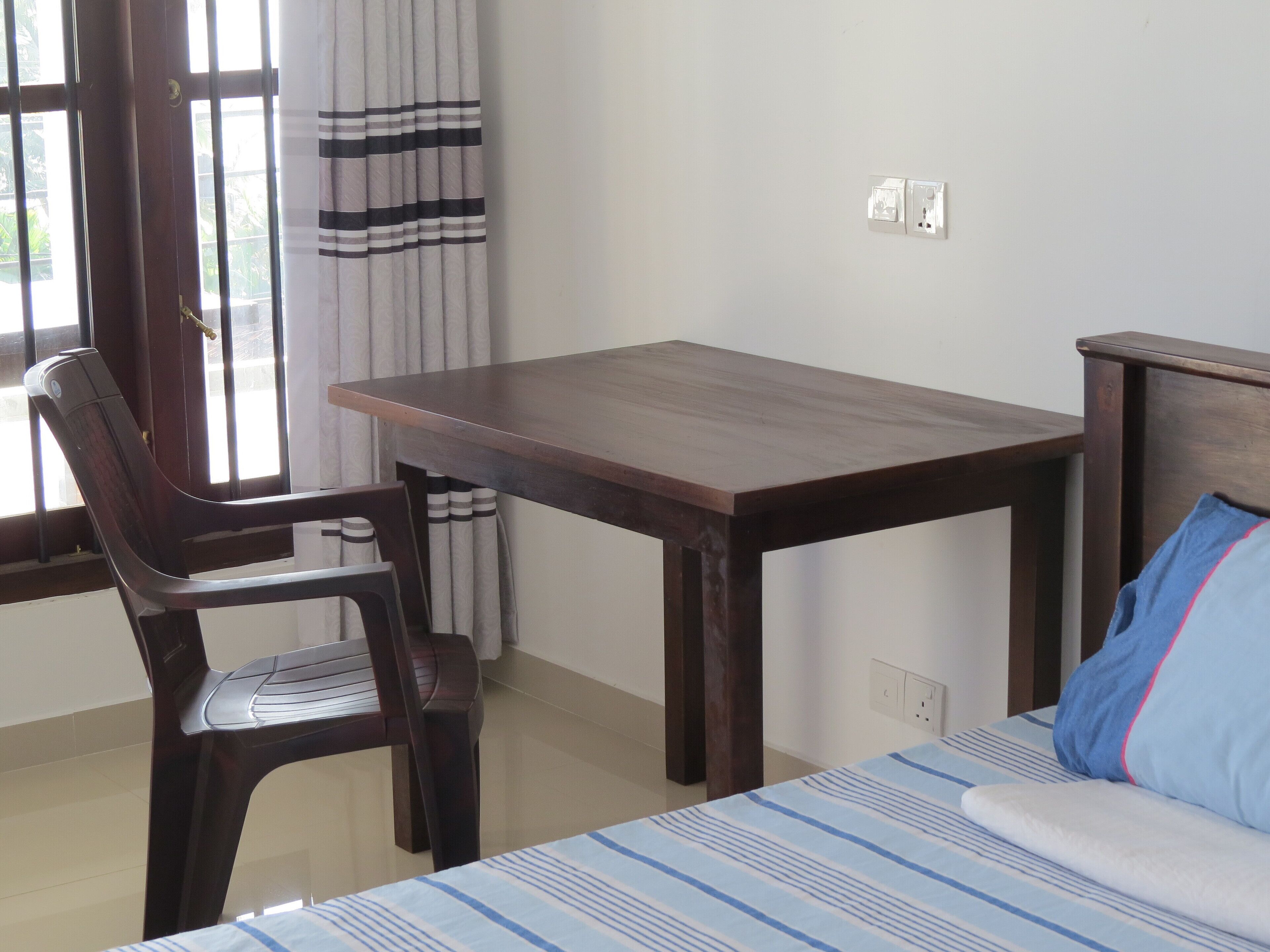 Une pièce avec un lit, une table en bois, une chaise en plastique et une fenêtre avec des rideaux.