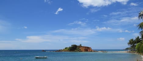 A beach with clear blue water, a small boat, and a rocky island with palm trees.