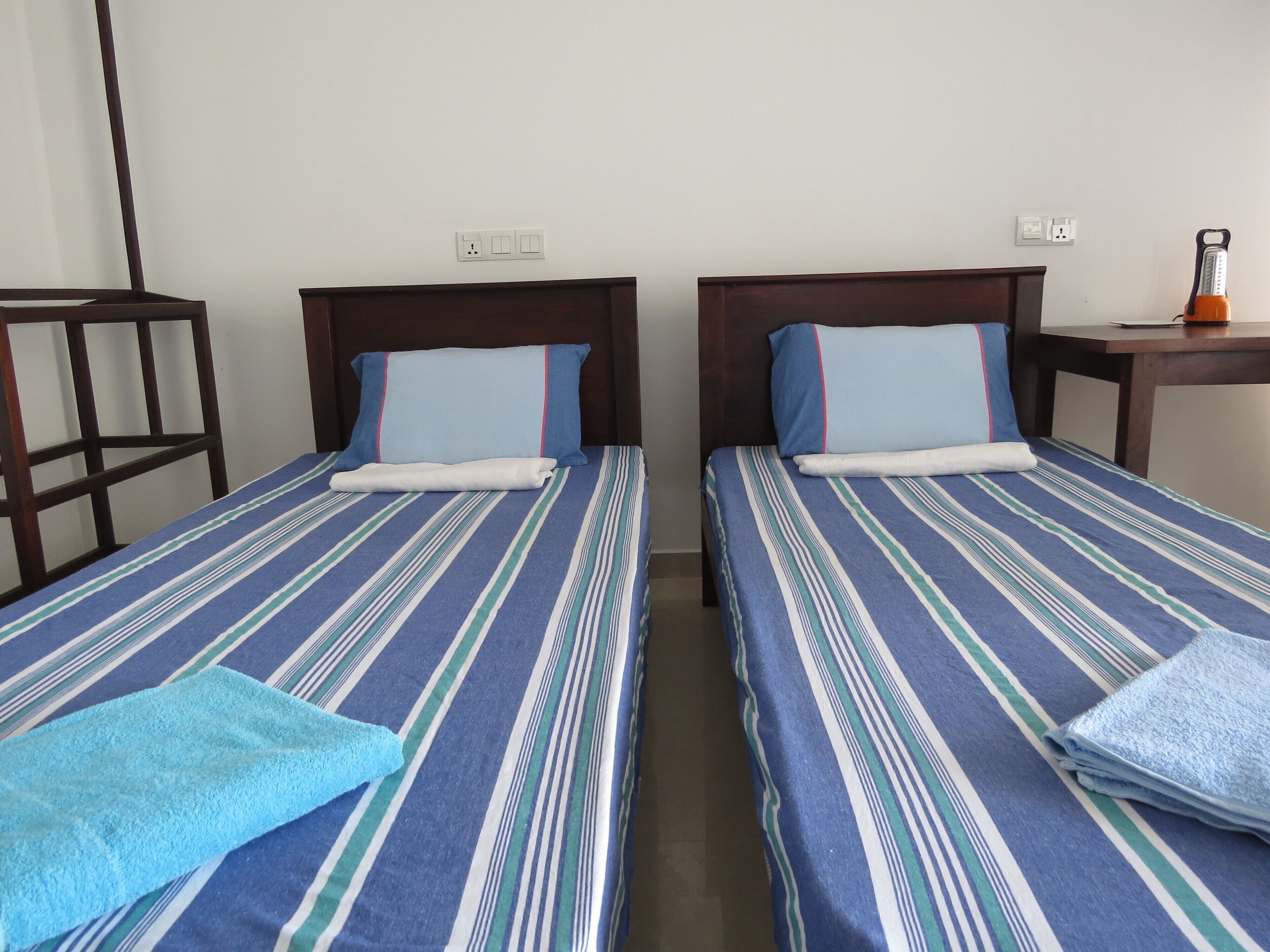 A hotel room with two single beds, each with striped bedding and pillows. There is a wooden headboard on each bed, and a small table with a lamp and a power outlet on the wall.
