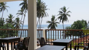 A balcony with wooden tables and chairs overlooking a beach with palm trees and the ocean. - Sailors Mirissa (Mirissa)