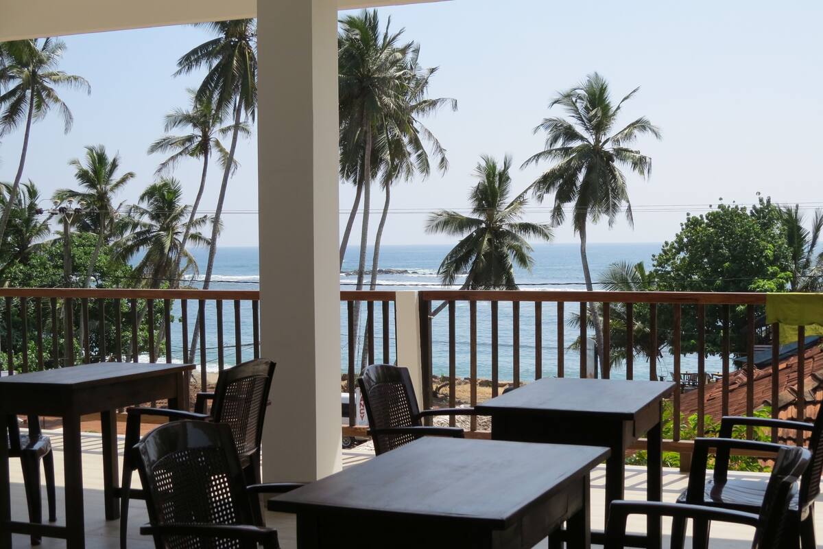 A balcony with wooden tables and chairs overlooking a beach with palm trees and the ocean.