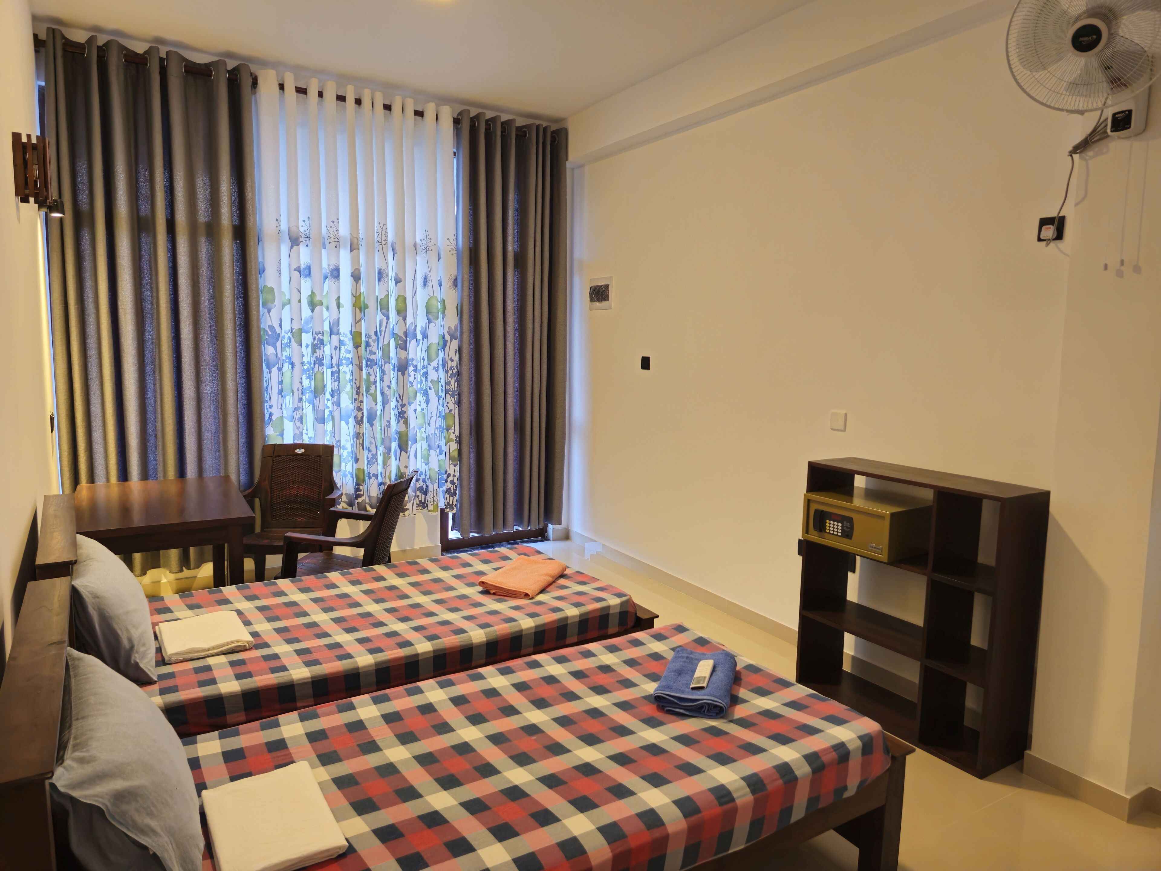 Una habitación de hotel con dos camas, un escritorio, una silla, un ventilador y un estante pequeño.