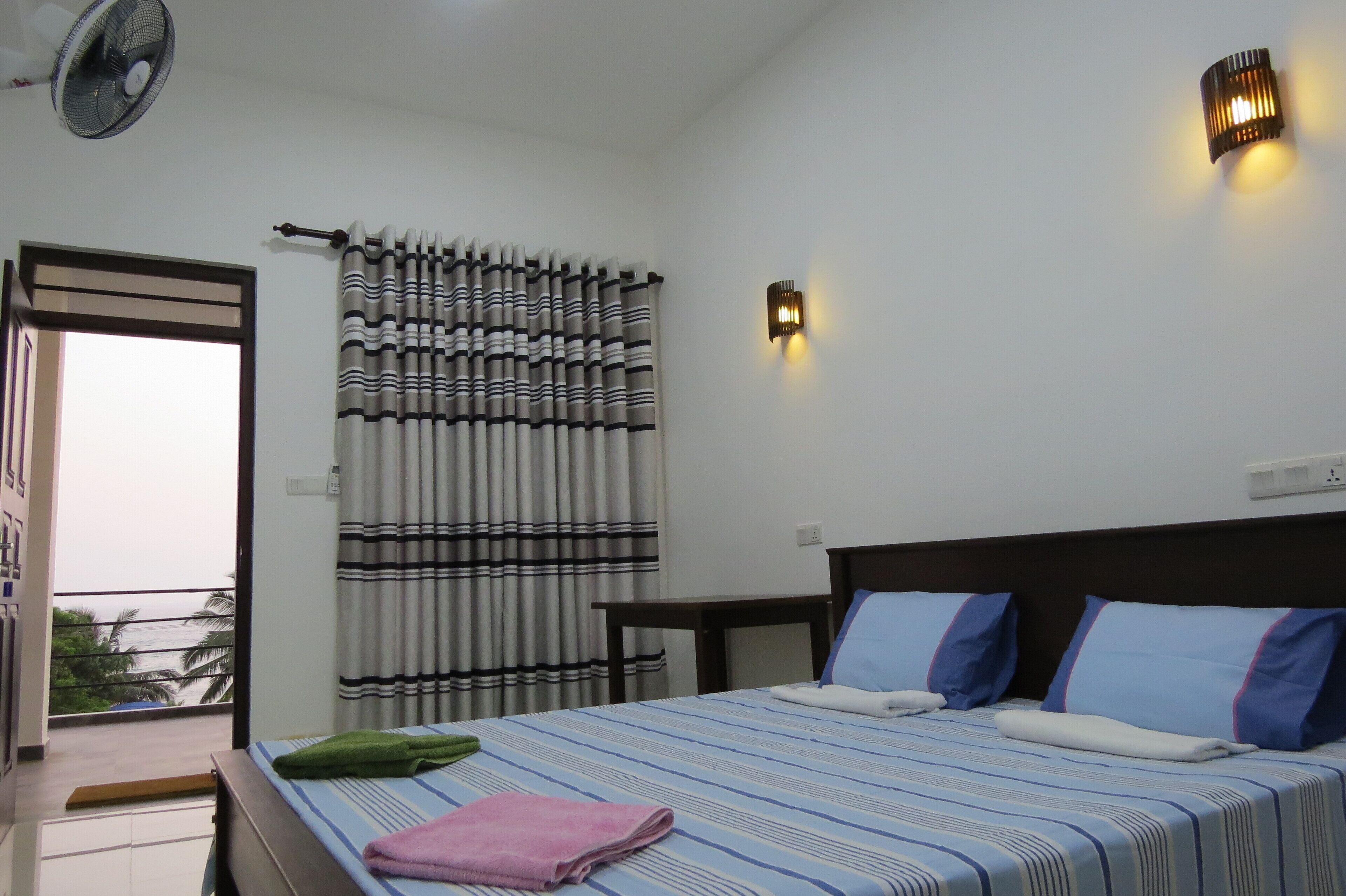 A hotel room with a bed, a bedside table, a wall-mounted light, a window with curtains, and a balcony with a view of palm trees.