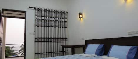 A hotel room with a bed, a bedside table, a wall-mounted light, a window with curtains, and a balcony with a view of palm trees.