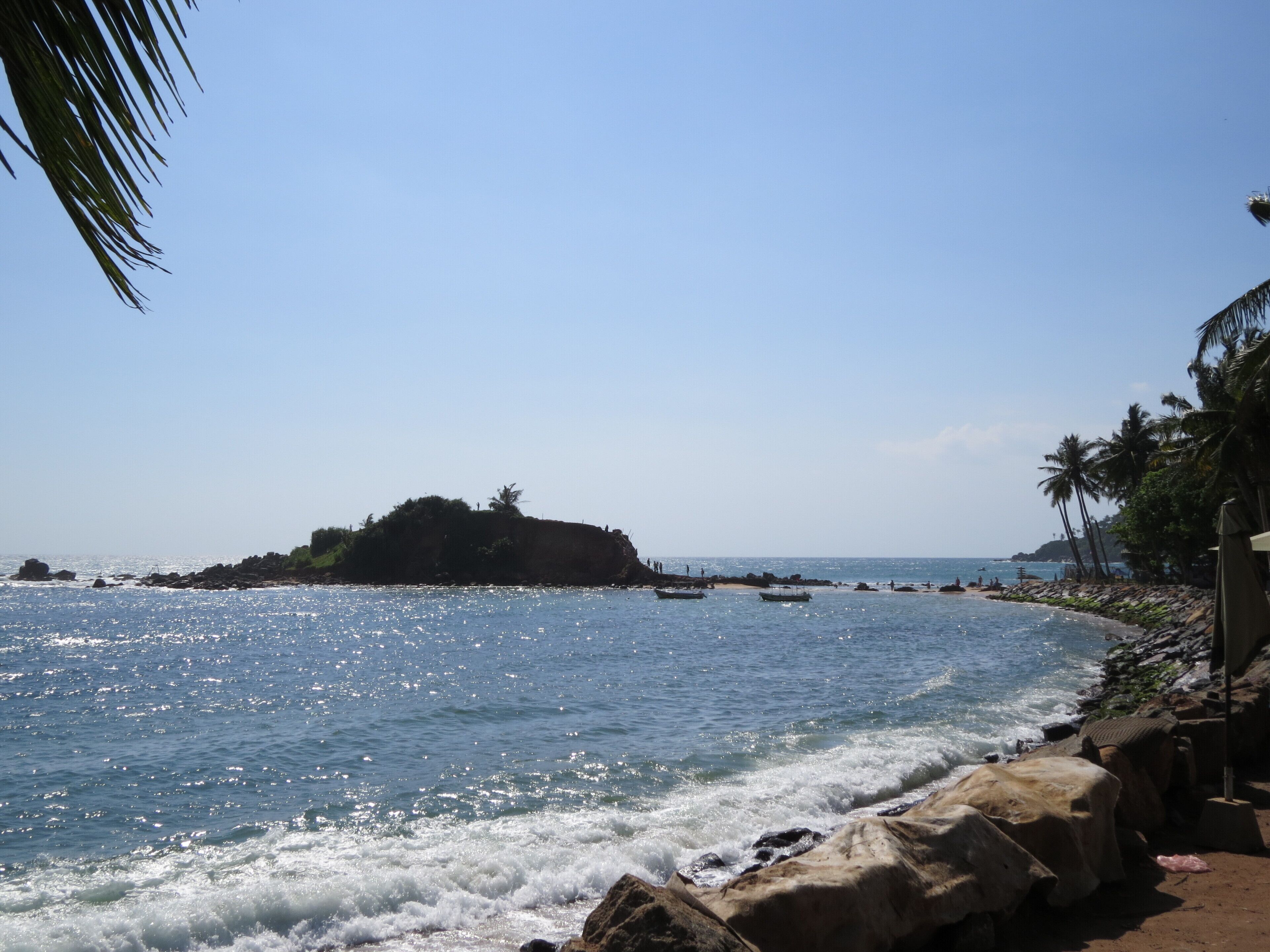 Uma praia com águas azul-claras, um afloramento rochoso e palmeiras.