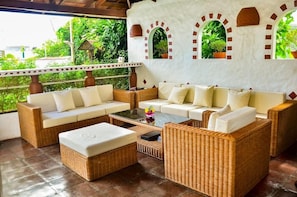 A covered outdoor seating area with wicker furniture and white cushions, surrounded by a white wall with arched windows and potted plants.