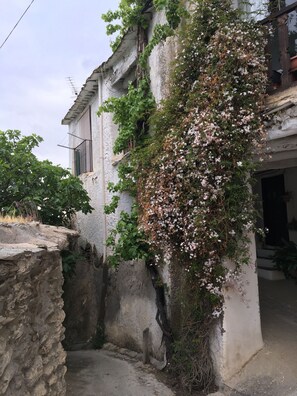 Un edificio con fachada de piedra, un pequeño balcón con barandilla metálica y un jardín con plantas en flor.