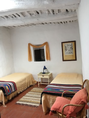 Une chambre avec deux lits, une petite table avec une lampe, une chaise, un tableau encadré au mur et une fenêtre avec des rideaux.
