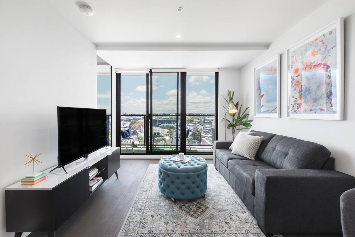 Un salón moderno con un televisor de pantalla plana grande, un sofá gris, un reposapiés azul y vistas de la ciudad a través de ventanales.