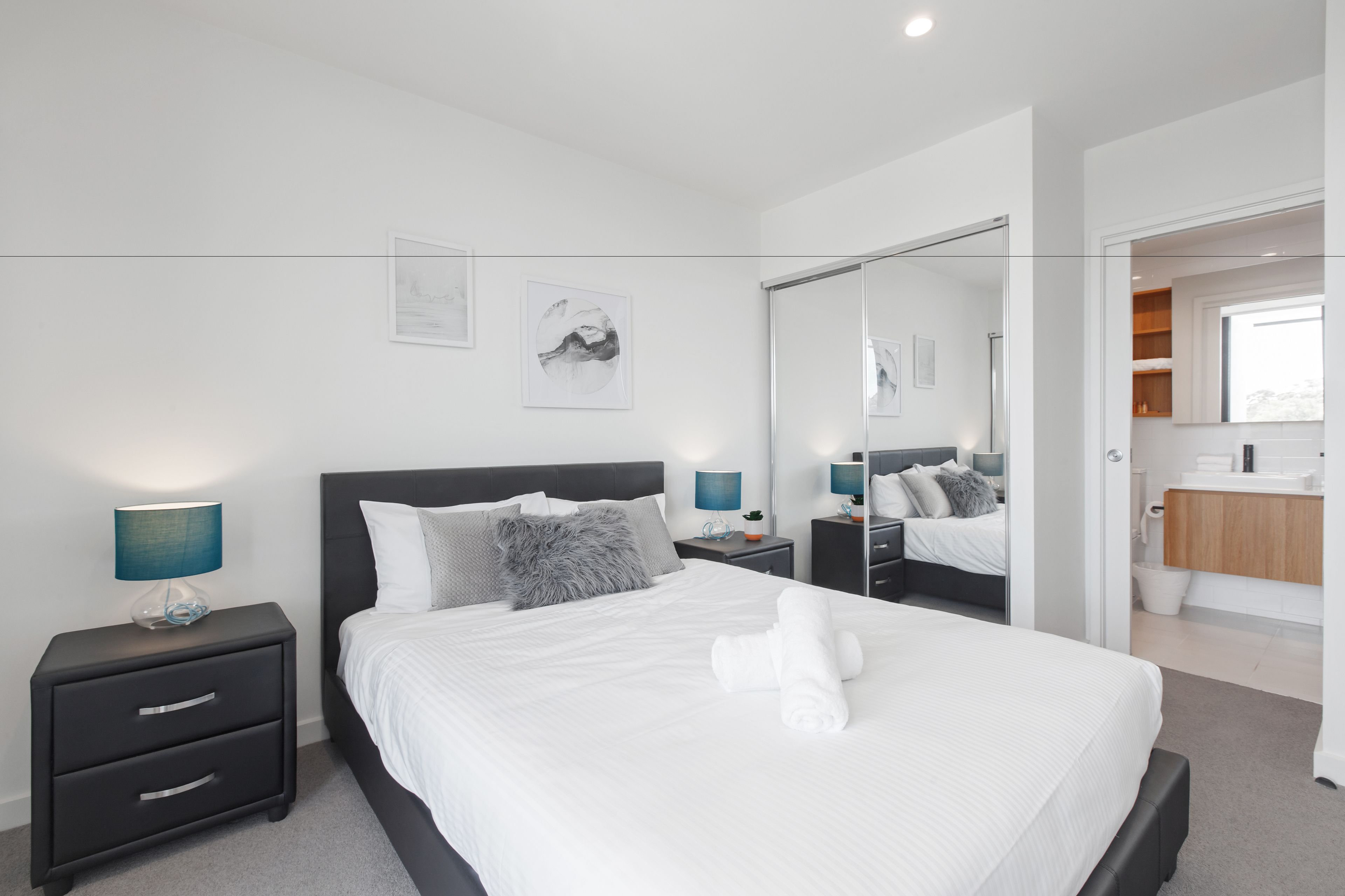 Une chambre d’hôtel moderne, dotée d’un grand lit, d’une table de chevet avec des tiroirs, d’une petite table de nuit avec une lampe, et d’une salle de bain visible à travers une porte ouverte.