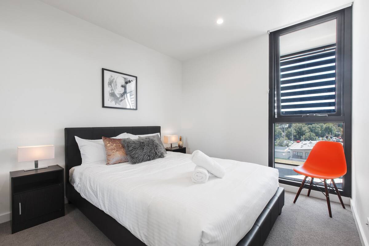 Un dormitorio moderno con una cama grande, una mesita de noche con lámpara, un cuadro en la pared y una ventana con persianas.