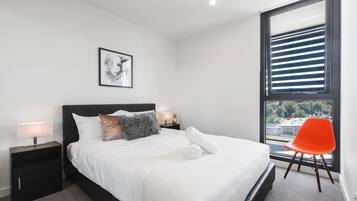 A modern bedroom with a large bed, a nightstand with a lamp, a framed picture on the wall, and a window with blinds.