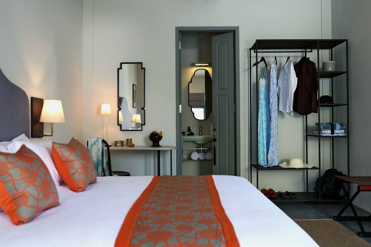Habitación de hotel con cama, escritorio con lámpara, espejo, armario y baño visible a través de una puerta abierta.