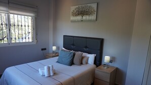 Een netjes opgemaakt bed met hoofdeinde, twee nachtkastjes met lampen, een raam met tralies en een schilderij aan de muur.