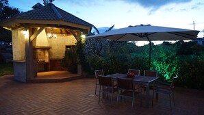 Een terras buiten met een prieeltje, een tafel en stoelen, en een parasol.
