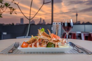 Un plato de mariscos sobre una vajilla blanca, acompañado de una copa de vino tinto, colocado en una mesa con vista a un horizonte urbano al atardecer.