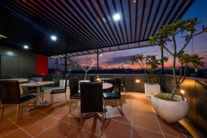 Un área de comedor al aire libre con mobiliario moderno, una planta grande en maceta y vista a la ciudad al atardecer.