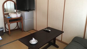 A compact living space with a dark wooden table, a sofa, a television on a stand, a small refrigerator, and a wicker chair.