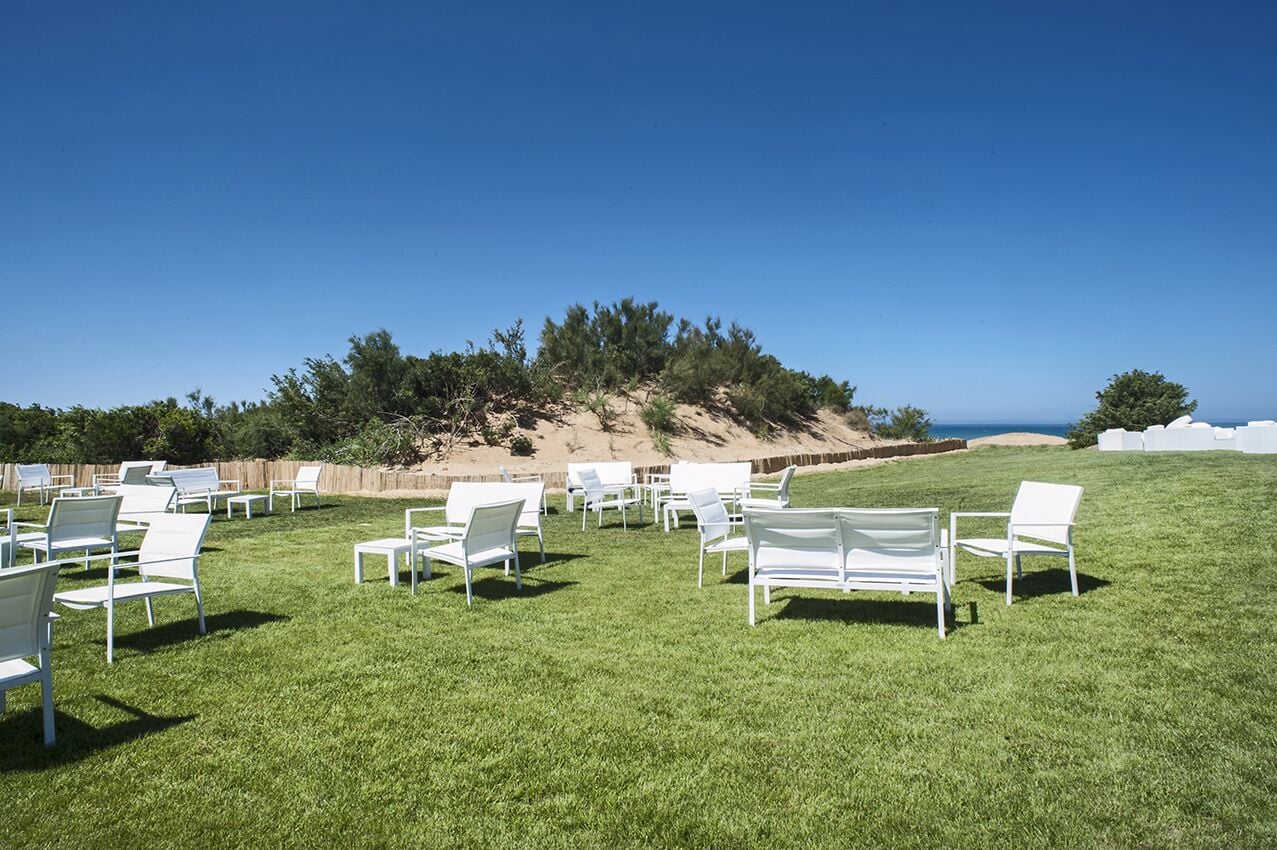 An outdoor seating area with white chairs and tables arranged on a grassy field, overlooking a beach and a sandy hill with trees.