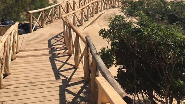 Un paseo marítimo de madera que lleva a una playa, con una vista despejada del océano y edificios a lo lejos.