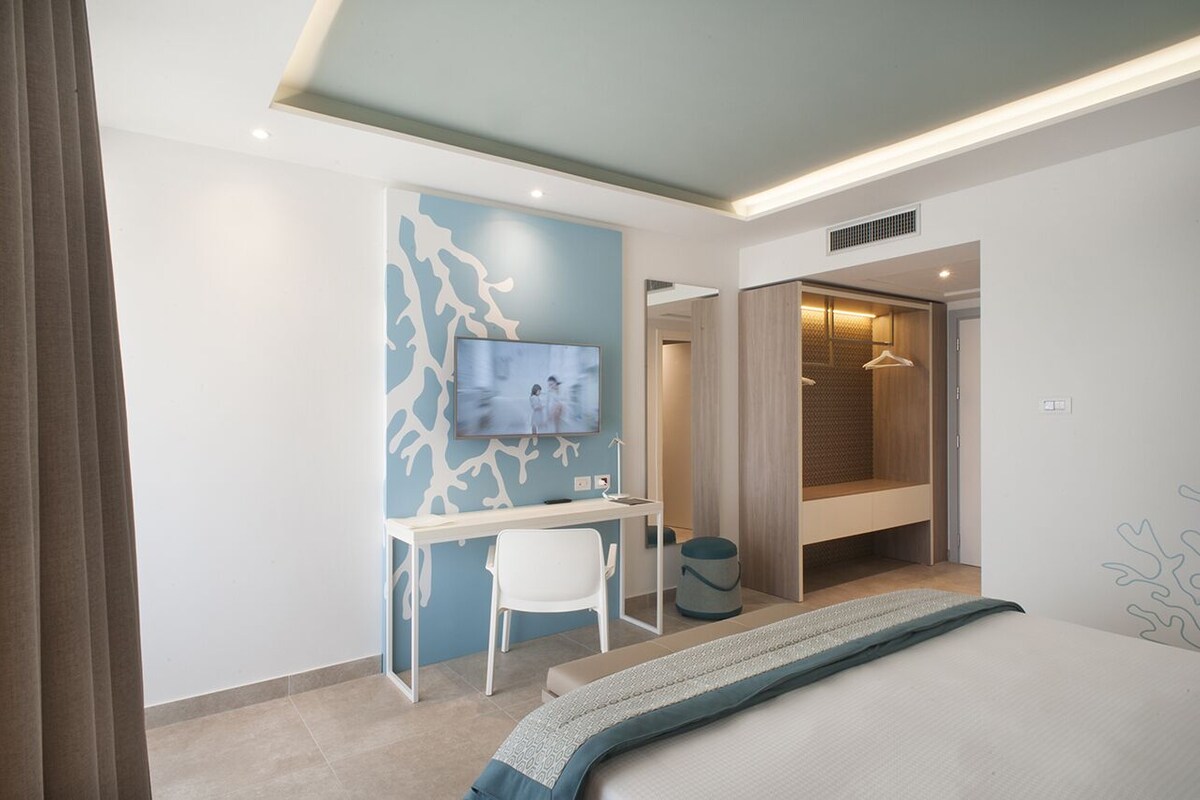 A modern hotel room with a bed, a desk with a chair, a television, and a walk-in closet.