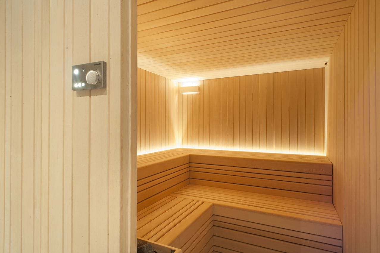 A sauna with wooden walls and benches, a control panel, and a light strip along the ceiling.
