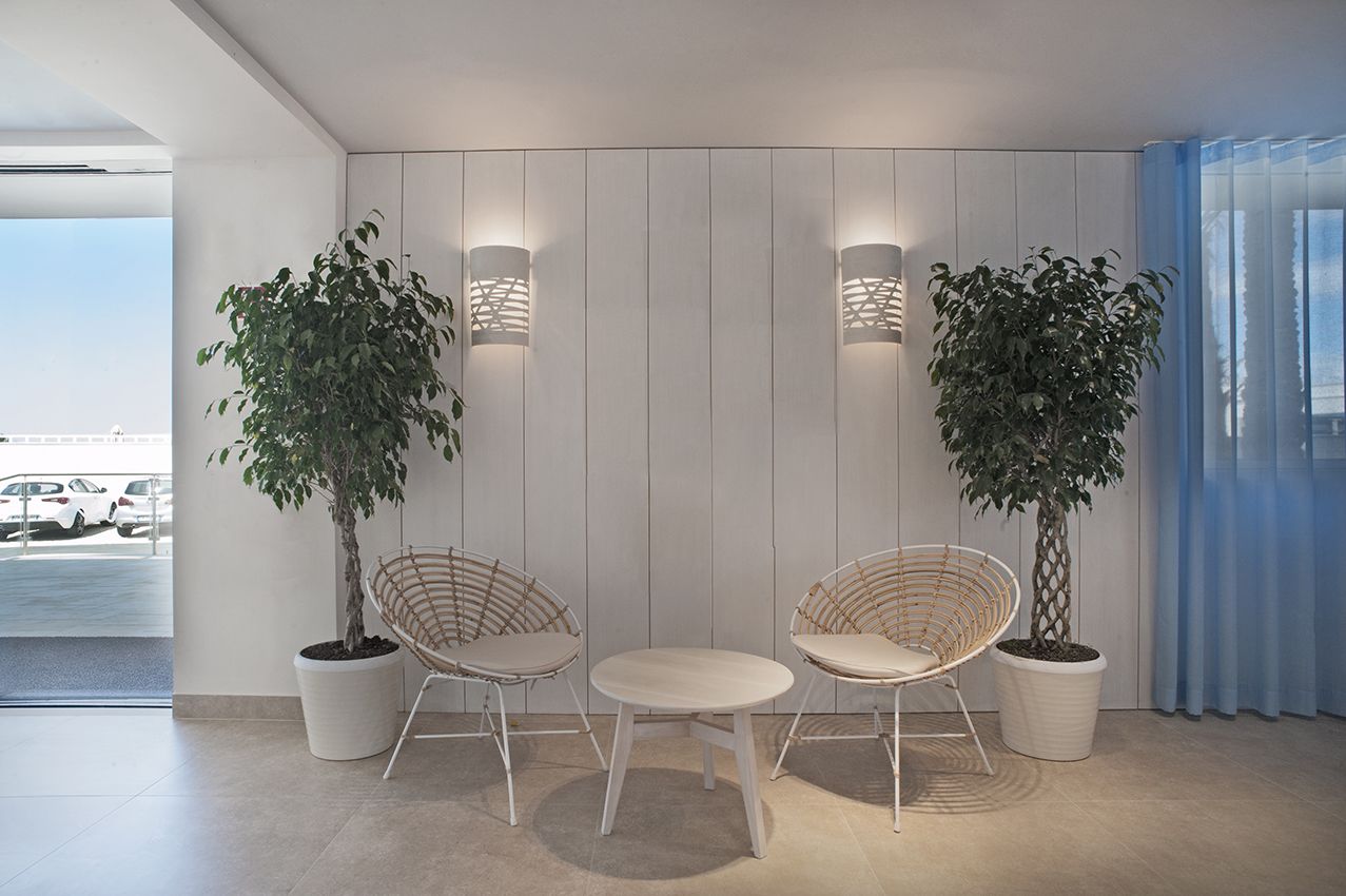 Een modern interieur met twee rieten stoelen en een kleine tafel, twee potplanten, en een uitzicht op een parkeerplaats door een glazen deur.