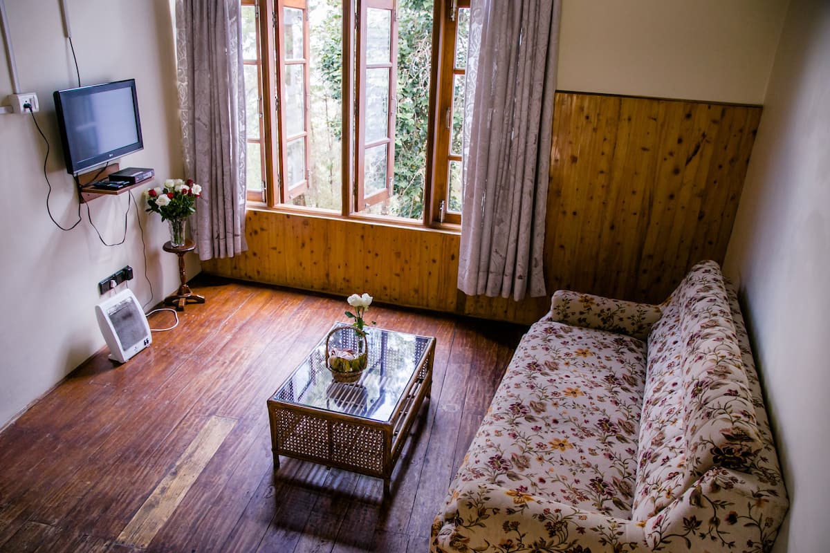 A room with a wooden floor, a sofa with floral upholstery, a glass coffee table with a vase of flowers, a television on a stand, and a window with curtains.