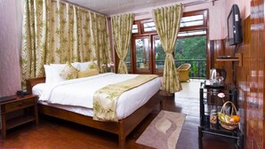 Une chambre avec un grand lit, une tête de lit en bois, une table de chevet, un téléviseur fixé au mur, une petite table avec une bouilloire et un panier de fruits, et un balcon avec une chaise.