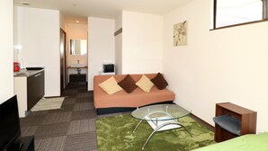 Habitació d'hotel compacta amb sofà, taula de centre de vidre, televisió, petita cuina i bany modest.