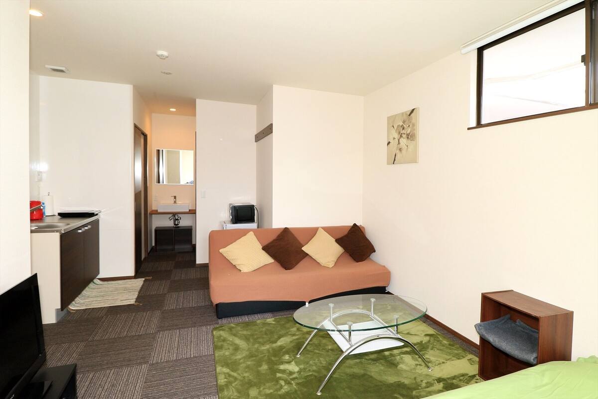 A compact hotel room with a sofa, a glass coffee table, a TV, a kitchenette, and a small bathroom.