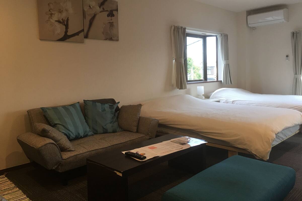 A hotel room with a sofa, a bed, a coffee table with a remote control, and two wall-mounted paintings.