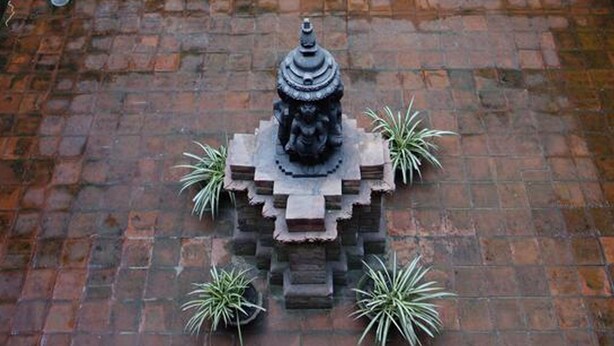 Una statua in pietra con tetto a pagoda, circondata da piante in vaso su un muro di mattoni.