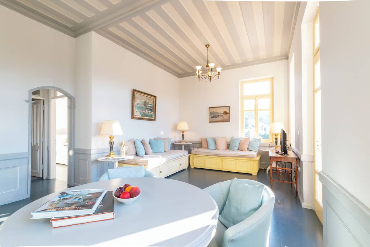 A well-lit room with a round table, a sofa, and a bed. There is a chandelier hanging from the ceiling, and a window providing natural light.