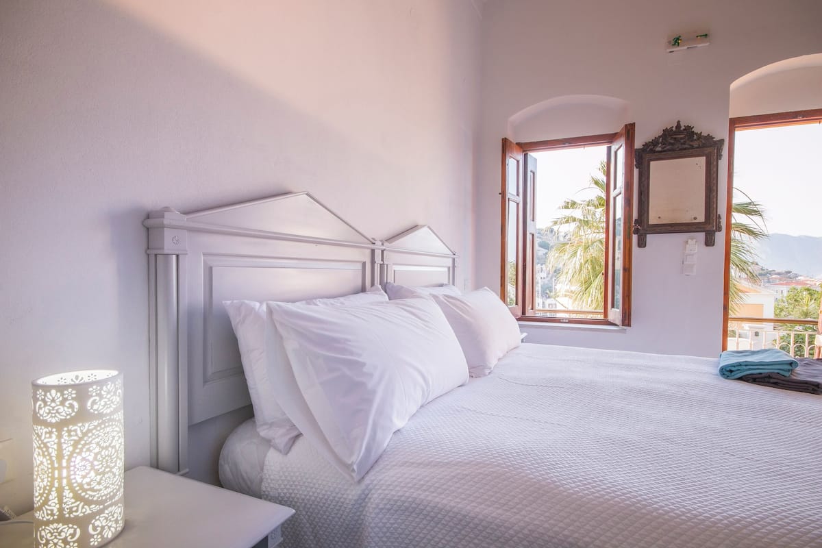 A bedroom with a large bed, a nightstand, a lamp, and a window with a view of palm trees and buildings.