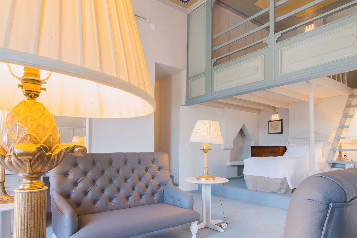 A room with a tufted sofa, a round table, a lamp, and a bed with a headboard.