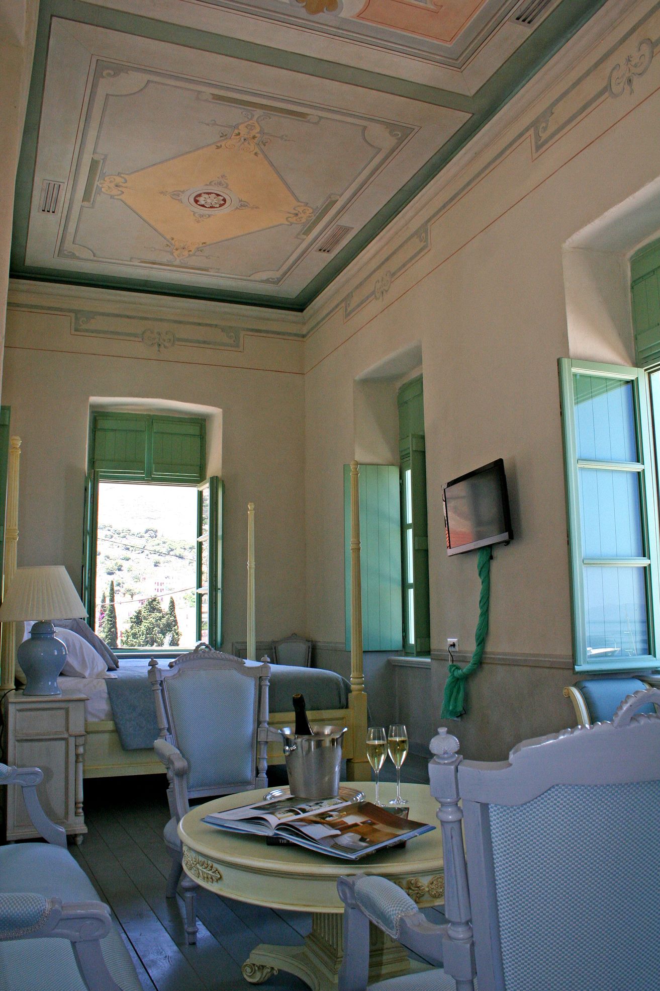 A room with a bed, a table with a magazine, a bottle, and two glasses of wine. There is a TV on the wall and a window with green shutters.