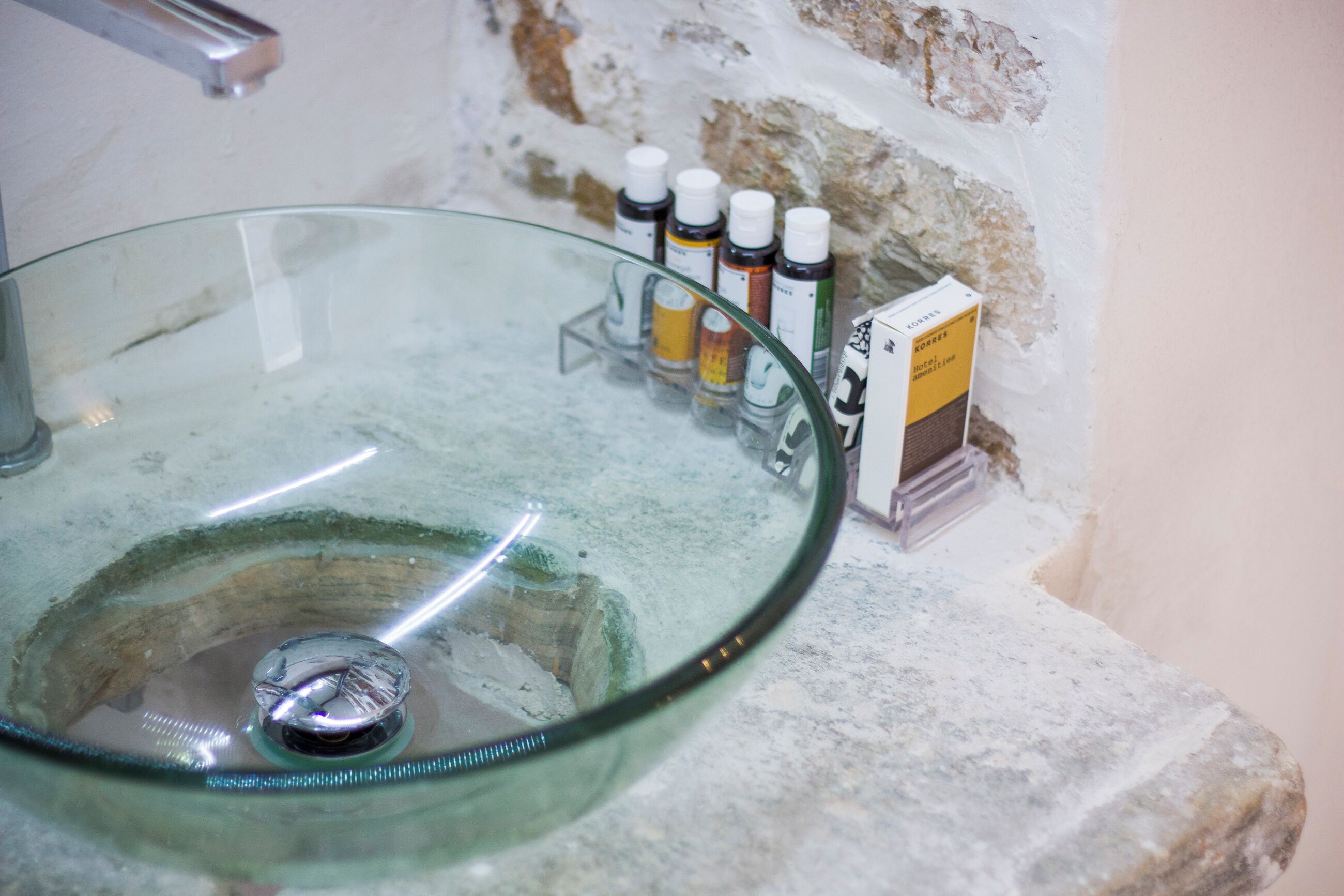 A glass sink with a metal drain, surrounded by a stone wall and a shelf with various bottles and a box.