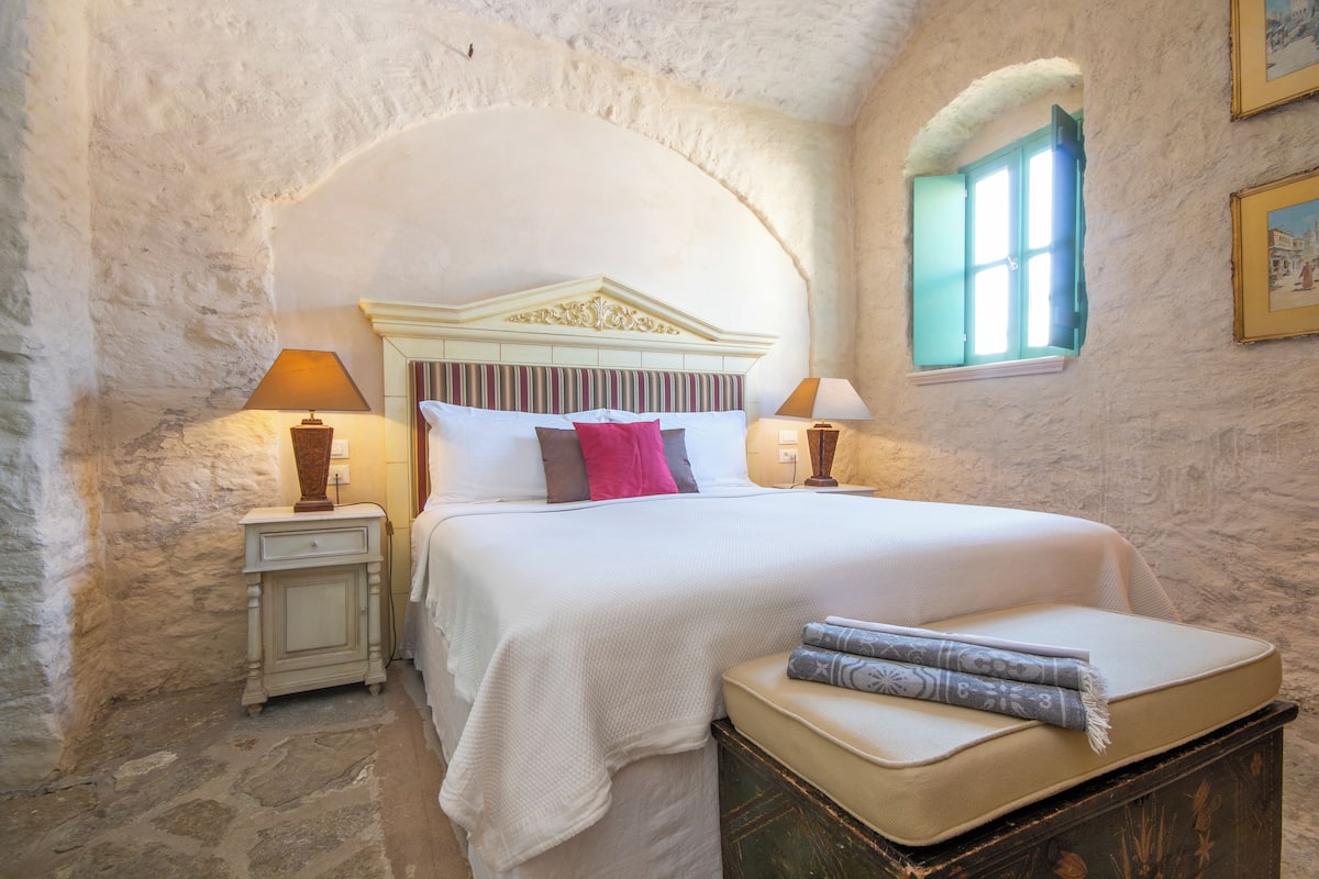 A bedroom with a stone wall, a bed with a headboard, two bedside tables with lamps, a window with blue shutters, and a small table with a cushion.