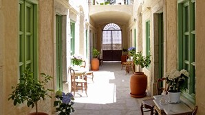 Un couloir étroit aux portes et fenêtres vertes, qui mène à un espace central où se trouvent une table et des chaises, le tout étant entouré de plantes en pot.