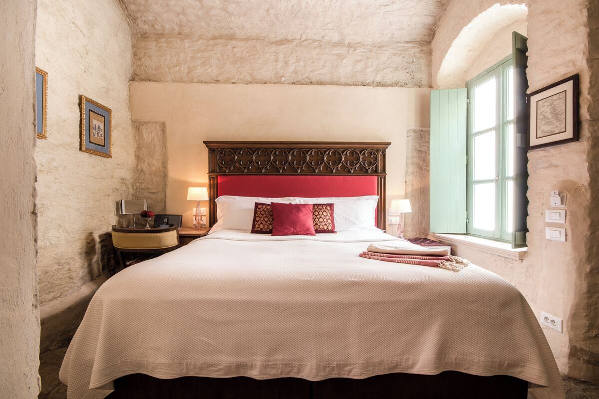 A bed with a red headboard, white bedding, and a window with green shutters.