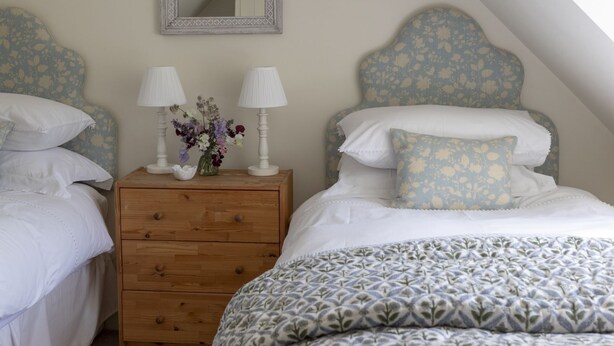 A bedroom with a sloped ceiling, a wooden nightstand with two lamps, a mirror, and a bed with patterned bedding. - GARDEN COTTAGE  - peaceful rural cottage in Scottish countryside (Denholm)