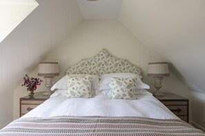 A bedroom with a sloped ceiling, a bed with white bedding and patterned pillows, two bedside lamps, and a nightstand with a vase of flowers. - GARDEN COTTAGE  - peaceful rural cottage in Scottish countryside (Denholm)