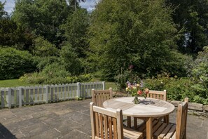 A wooden table and chairs set on a stone patio with a view of a lush garden and a wooden fence.