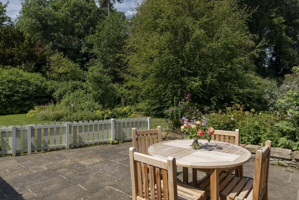 A wooden table and chairs set on a stone patio with a view of a lush garden and a wooden fence. - GARDEN COTTAGE - peaceful rural cottage in Scottish countryside (Denholm)