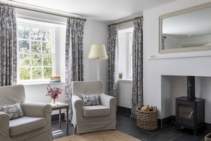 A cozy living room with a fireplace, checkered armchairs, a lamp, and a view of greenery outside.