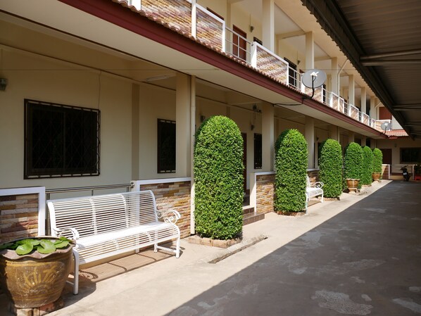 A covered outdoor area with a row of potted plants, a white bench, and a building with multiple windows and a balcony.