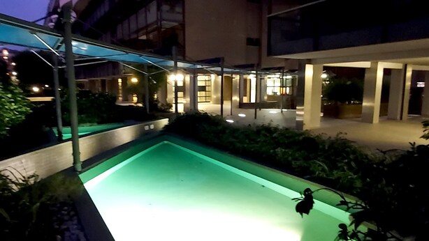 An outdoor swimming pool area at dusk with a covered seating area and a building in the background.